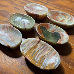 May include: Six oval-shaped, decorative bowls made of natural onyx stone. The bowls have a variety of colors and patterns, including white, brown, green, and red.