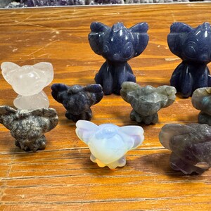 May include: A collection of small, carved stone figurines in various colors, including blue, white, and gray. The figurines are in the shape of a cartoon character. The figurines are displayed on a wooden surface.