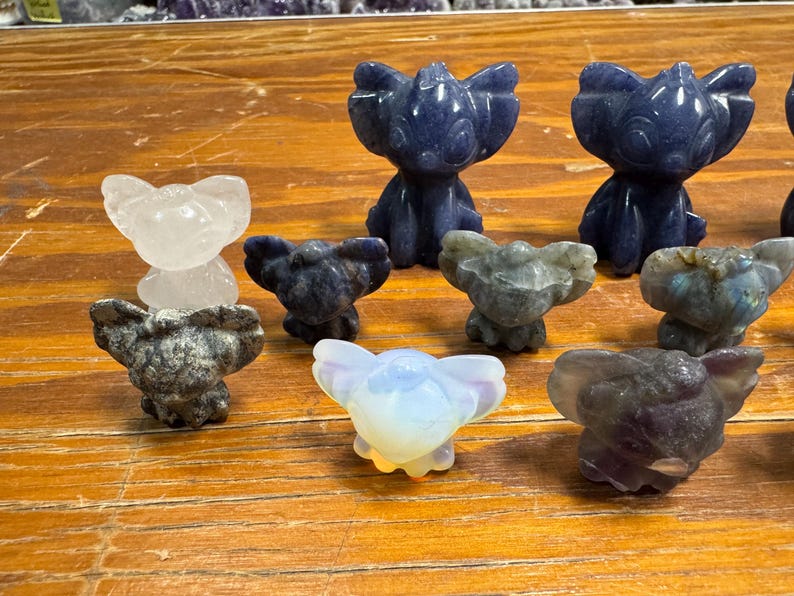 May include: A collection of small, carved stone figurines in various colors, including blue, white, and gray. The figurines are in the shape of a cartoon character. The figurines are displayed on a wooden surface.