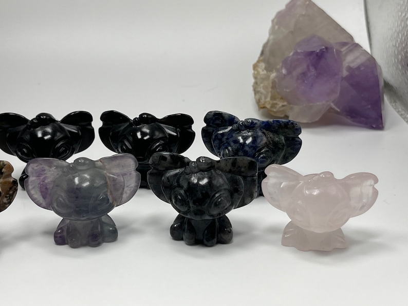 May include: A collection of carved stone figurines in various colors, including black, blue, purple, and pink. The figurines are in the shape of a cartoon character. A large amethyst crystal is in the background.