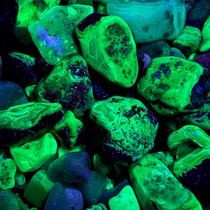 Puede incluir: Un conjunto de rocas brillantes de color verde y azul. Las piedras tienen varias formas y tamaños, algunas con patrones intrincados. La imagen oscura resalta los colores fluorescentes vibrantes de las rocas.