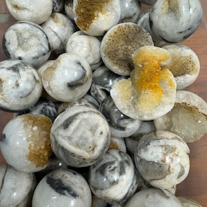 May include: A collection of white and gray geode spheres with a crystal interior. The spheres have a natural, rough texture and are arranged in a pile.