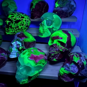 May include: A collection of skull-shaped sculptures featuring a mix of dark maroon and vibrant neon green colors. These objects exhibit a marbled or swirled pattern and glow intensely under ultraviolet light, highlighting their detailed anatomical features.