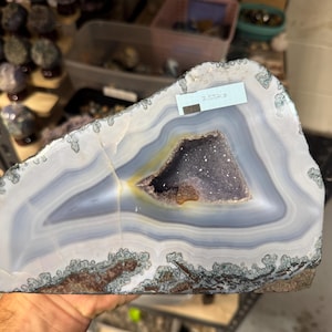 May include: A large, polished agate slice featuring concentric bands of white, gray, and translucent blue, with a central geode cavity containing dark purple crystals. A white label on the top edge displays the text 3.55kg.