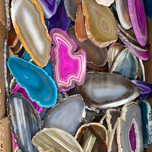 May include: A collection of agate slices in various colors and patterns, including pink, blue, purple, brown, and gray. The slices are arranged in a box, with some overlapping.