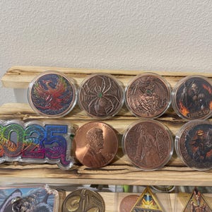 May include: A collection of collectible coins and tokens displayed on a wooden shelf. The coins feature various designs, including a phoenix, spider, and a figure with the text "TIMES UP". A 2025 token is also visible.