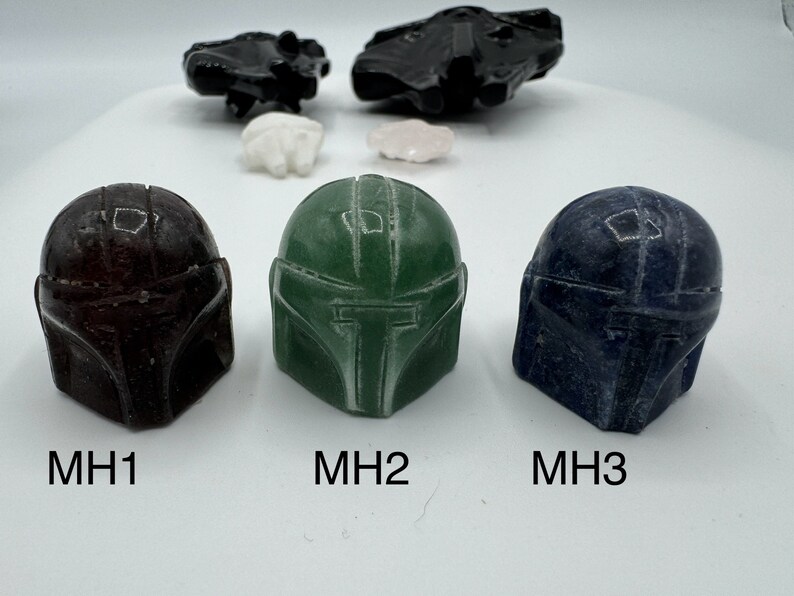 May include: Three Mandalorian helmet-shaped resin figurines. The helmets are in different colors: dark brown, green, and blue. The helmets are labeled MH1, MH2, and MH3.
