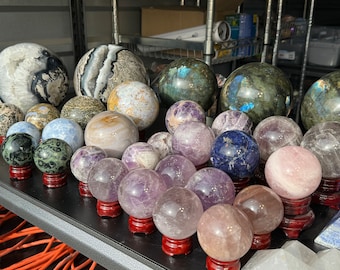 May include: A collection of various polished gemstone spheres on wooden stands. The spheres are made of amethyst, rose quartz, labradorite, agate, and other types of stones. The spheres are arranged in a semi-circle on a black surface.