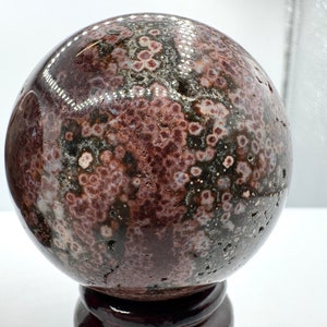 May include: A polished, spherical gemstone with a speckled pattern of maroon, gray, and black. The orb is displayed on a dark wooden stand, showcasing its natural beauty. The stone's surface has a glossy finish.