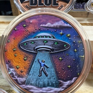 May include: A round, copper-colored coin featuring a detailed image of a person being abducted by a UFO. The background depicts a colorful night sky with stars and clouds. The coin is encased in a clear protective case.
