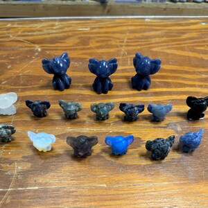 May include: A collection of small, carved elephant figurines in various colors, including blue, black, and clear. The elephants are arranged on a wooden surface, showcasing their detailed features and unique stone textures.