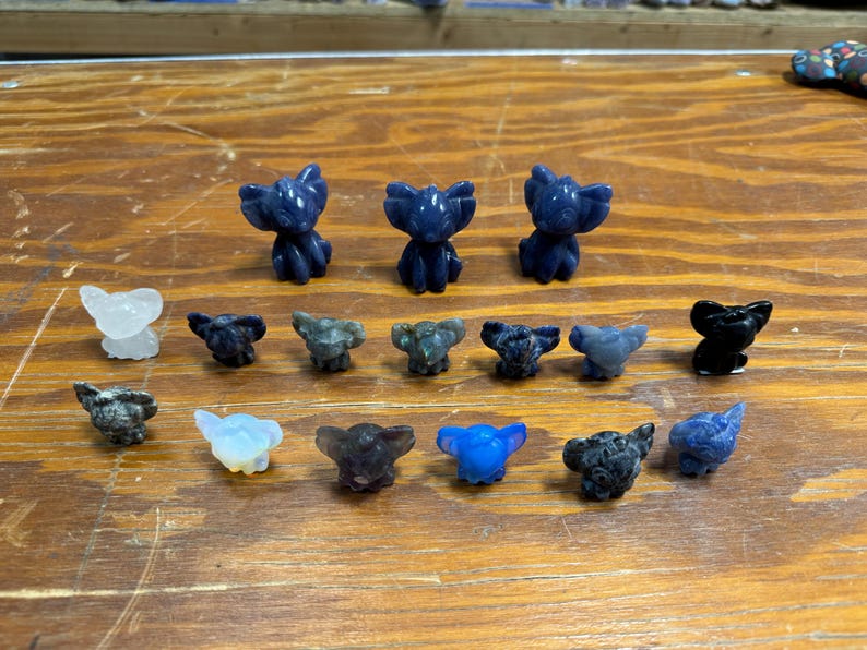May include: A collection of small, carved elephant figurines in various colors, including blue, black, and clear. The elephants are arranged on a wooden surface, showcasing their detailed features and unique stone textures.