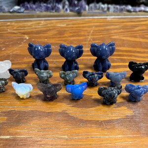 May include: A collection of carved stone figurines in various colors, including blue, black, and white. The figurines are in the shape of a stylized animal, possibly a bear or a koala. They are arranged on a wooden surface.