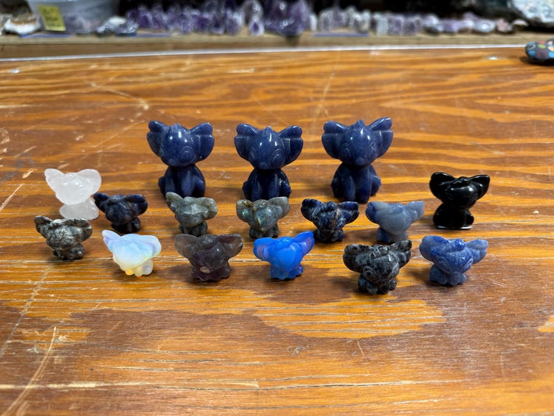 May include: A collection of carved stone figurines in various colors, including blue, black, and white. The figurines are in the shape of a stylized animal, possibly a bear or a koala. They are arranged on a wooden surface.