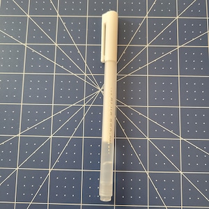 May include: A white gel pen with a cap and clip, lying on a blue cutting mat with a grid pattern. The pen has text printed vertically along its side. The pen is a writing tool.