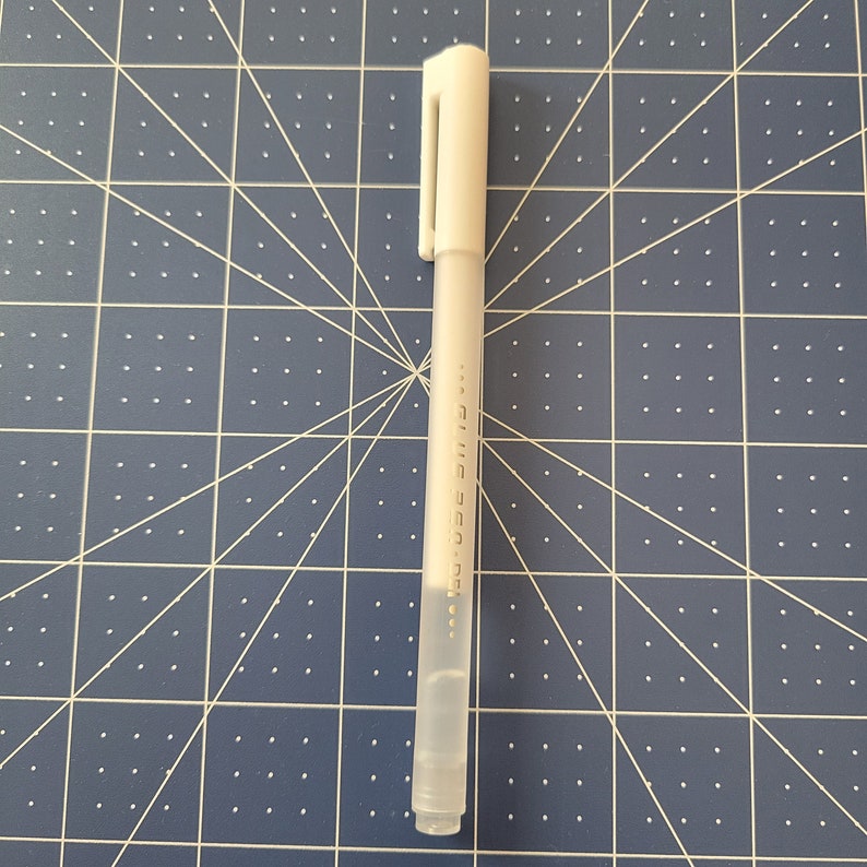 May include: A white gel pen with a cap and clip, lying on a blue cutting mat with a grid pattern. The pen has text printed vertically along its side. The pen is a writing tool.