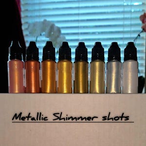 May include: A row of small dropper bottles filled with metallic shimmer shots in various colors, including rose gold, copper, and gold. The bottles have black caps and are labeled "Metallic Shimmer shots".