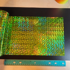 Puede incluir: Una lámina de vinilo holográfico verde iridiscente con un patrón geométrico. El vinilo tiene una superficie brillante y reflectante.