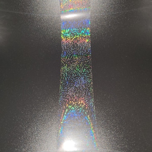 May include: A holographic transfer foil with a rainbow iridescent finish. The foil features a repeating pattern of stylized fireworks.