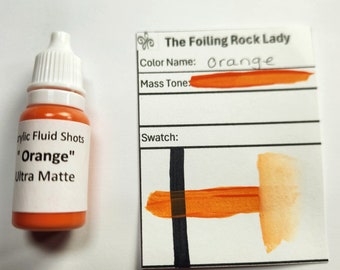 Puede incluir: Una pequeña botella de pintura acrílica naranja etiquetada "Acrylic Fluid Shots "Orange" Ultra Matte". Una tarjeta de muestra muestra el color de la pintura, etiquetada "The Foiling Rock Lady" y "Orange".
