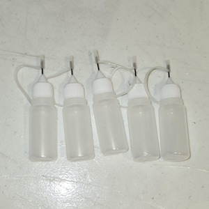Op de afbeelding: Vijf doorzichtige plastic flessen met witte doppen en doseerpunten zijn in een rij gerangschikt. Elke fles heeft een kleine, gebogen plastic lus aan de dop. De flessen staan op een wit oppervlak.