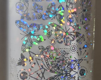 May include: A holographic sticker sheet with a repeating pattern of butterflies and floral designs. The sticker sheet has a silver background with iridescent colors.