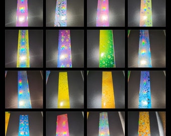 May include: A collection of iridescent, holographic tape strips in various colors, including pink, yellow, green, and blue. Some strips feature a water droplet design, while others are solid. The tape is displayed against a dark background.