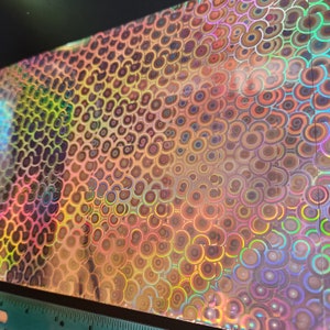 Op de afbeelding: Een vel iriserende holografische vinyl met een herhalend patroon van concentrische cirkels in tinten roze, oranje, geel, groen, blauw en paars.