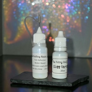 May include: Two small, white bottles of craft supplies on a dark gray surface. One bottle has a needle applicator and the other has a label that reads "Gloss Varnish .33oz". The other bottle reads "Foil Adhesive Water Based".