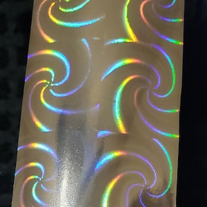 May include: A close-up of a silver holographic sheet with a repeating pattern of iridescent swirls. The swirls are a mix of blue, green, yellow, and purple.