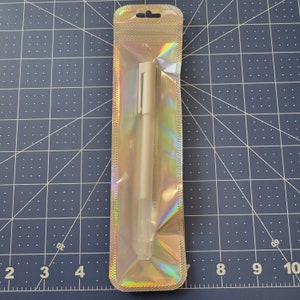 May include: A single white pen in a clear, iridescent plastic package. The pen has a simple design with a cap and a clip. The packaging has a small hole at the top for hanging. The background is a blue cutting mat with a grid pattern.