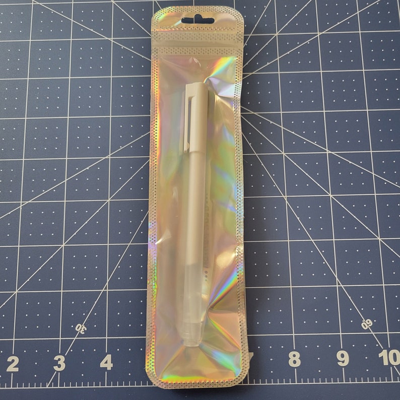May include: A single white pen in a clear, iridescent plastic package. The pen has a simple design with a cap and a clip. The packaging has a small hole at the top for hanging. The background is a blue cutting mat with a grid pattern.