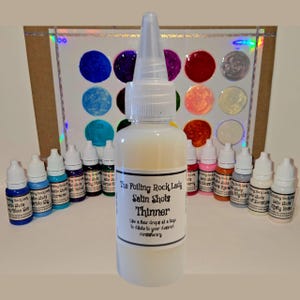 May include: A collection of art supplies featuring a bottle of "Satin Shots Thinner" and several small bottles of colorful paints. A color chart with various paint swatches is visible in the background. The text "The Foiling Rock Lady" is on the bottle.