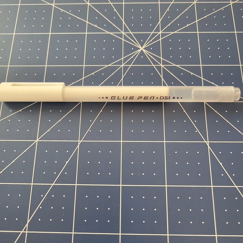 May include: A white glue pen with a clear tip and a rectangular cap. The text "... GLUE PEN &bull; D51 ..." is printed on the pen's barrel. The pen is lying on a blue cutting mat with a grid pattern.