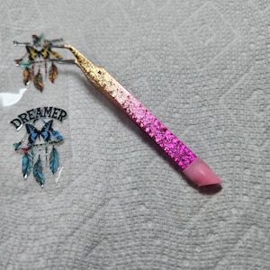 May include: A pink and gold glitter tweezer with a pink rubber tip. The tweezer is on a white textured surface.