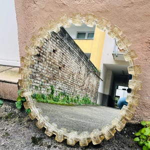 Può includere: Uno specchio rotondo con una cornice decorativa composta da blocchi di vetro rettangolari trasparenti. Lo specchio riflette un muro di mattoni e un edificio con pareti gialle e bianche. La cornice è di colore ambra chiaro.