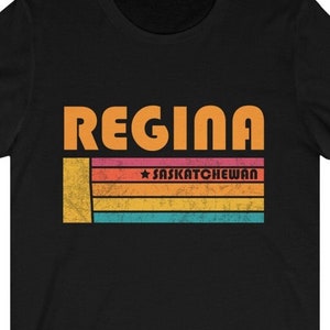 May include: Black t-shirt with the word "REGINA" in orange block letters above a horizontal striped design in orange, pink, yellow, and blue. The text "SASKATCHEWAN" is in the center of the stripes.
