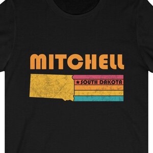 Könnte beinhalten: Schwarzes T-Shirt mit einer Grafik im Vintage-Stil des Bundesstaates South Dakota mit dem Text "MITCHELL" und "SOUTH DAKOTA" in Orange.