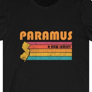 以下が含まれることがあります： オレンジ色の「PARAMUS, NEW JERSEY」の文字が入った、ニュージャージー州のヴィンテージスタイルのグラフィックがプリントされた黒いTシャツです。