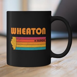 May include: A black ceramic mug featuring the word "WHEATON" in orange. Below is a yellow outline of Illinois, with horizontal stripes in orange, red, and blue. The word "ILLINOIS" is in red, in a retro style.