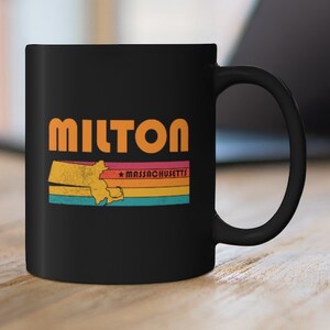 May include: Black coffee mug with a retro design featuring the word "MILTON" in orange text and a silhouette of the state of Massachusetts with the text "MASSACHUSETTS" in white. The mug has a white handle.