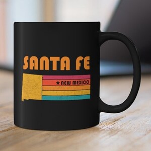 May include: Black ceramic coffee mug with a retro design featuring the text "SANTA FE" and a stylized outline of the state of New Mexico with the text "NEW MEXICO" below.