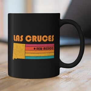 May include: Black coffee mug with a retro design featuring the text "LAS CRUCES" and a stylized outline of the state of New Mexico with the text "NEW MEXICO" below.
