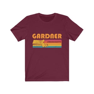 May include: A maroon t-shirt with a vintage-style graphic of the state of Massachusetts with the word "GARDNER" and "MASSACHUSETTS" in orange text.