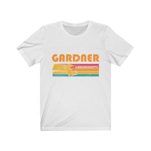 May include: White t-shirt with a vintage-style graphic of the state of Massachusetts with the word "GARDNER" and "MASSACHUSETTS" in orange text.