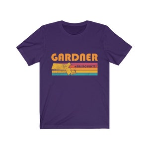 May include: Purple short-sleeved t-shirt with a vintage-style graphic of the state of Massachusetts with the word "GARDNER" and "MASSACHUSETTS" in orange text.