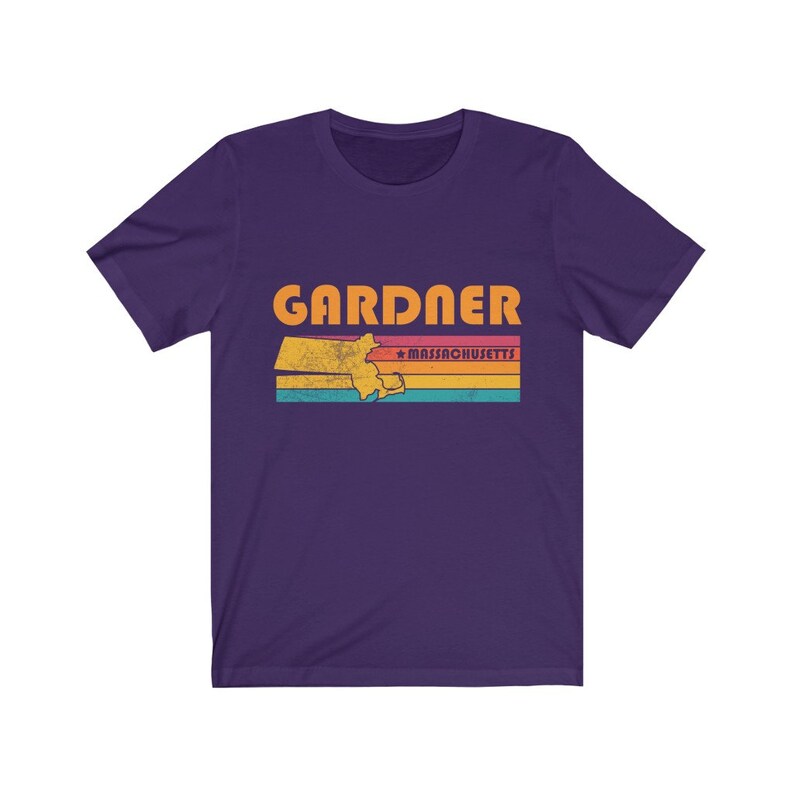 May include: Purple short-sleeved t-shirt with a vintage-style graphic of the state of Massachusetts with the word "GARDNER" and "MASSACHUSETTS" in orange text.