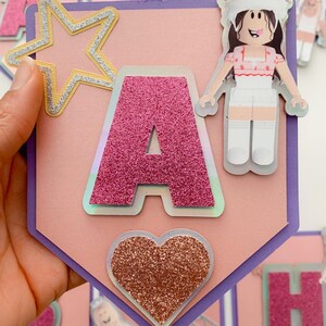 Puede incluir: Una pancarta rosa y morada con la letra A en purpurina rosa, una estrella dorada, un coraz&oacute;n rosa con purpurina y un personaje de dibujos animados con un vestido rosa y blanco. La pancarta dice "A"