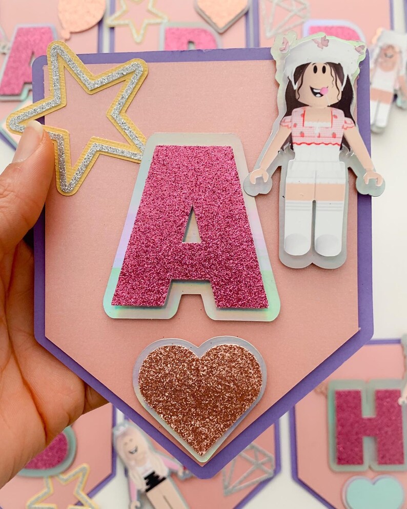 Puede incluir: Una pancarta rosa y morada con la letra A en purpurina rosa, una estrella dorada, un coraz&oacute;n rosa con purpurina y un personaje de dibujos animados con un vestido rosa y blanco. La pancarta dice "A"