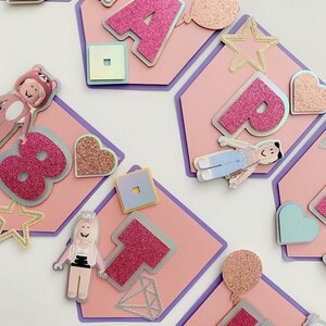 Puede incluir: Bandera rosa y morada con la palabra "Happy" en letras de purpurina rosa. Cada letra est&aacute; en una tarjeta separada con forma de hex&aacute;gono con varias decoraciones, incluyendo estrellas, corazones y un personaje de Roblox.